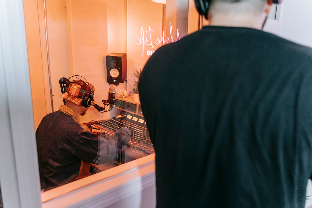 Two individuals in a recording studio using audio equipment, viewed through window.
